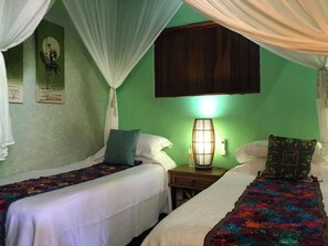 A room with two single beds, each with a white bedspread and a colorful floral blanket. There is a nightstand with a lamp between the beds, and a wooden cabinet above the nightstand. The walls are painted green, and there are two framed artworks on the wall.
