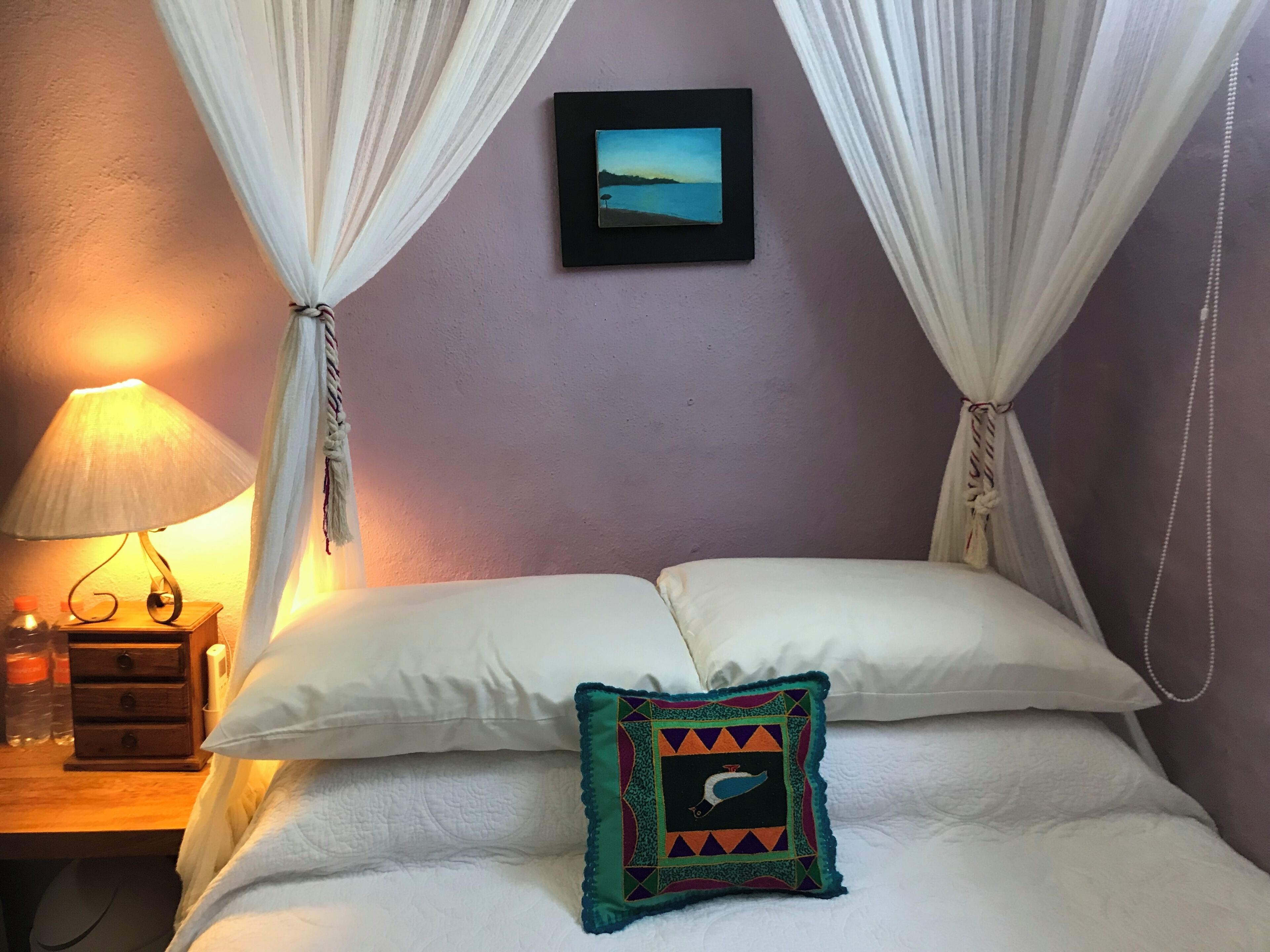 Un lit avec du linge de lit blanc, un coussin décoratif, un tableau accroché au mur et une lampe de chevet.