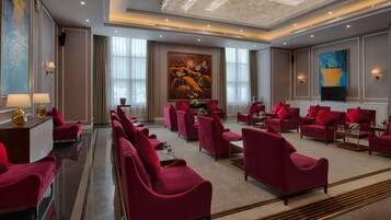 A spacious lobby with red upholstered sofas arranged in rows, a large painting on the wall, and a modern chandelier.