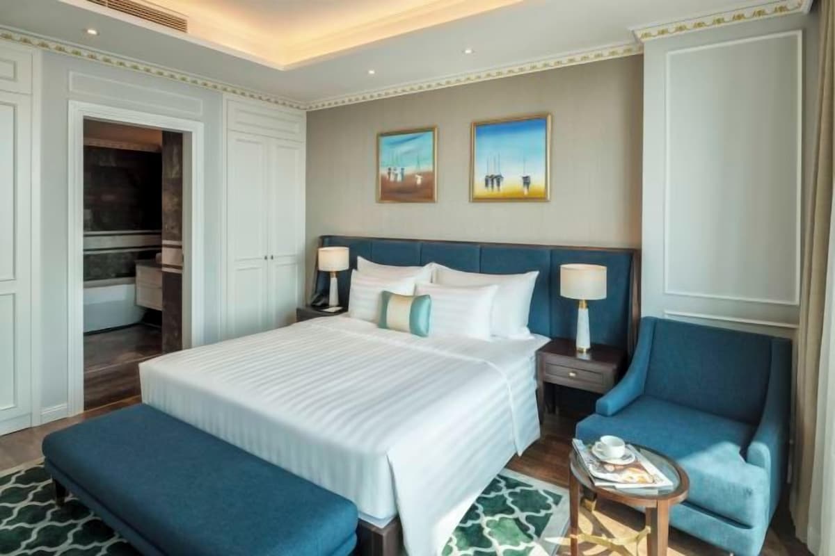 A hotel room with a large bed, two nightstands, a blue armchair, and a small table with a lamp.
