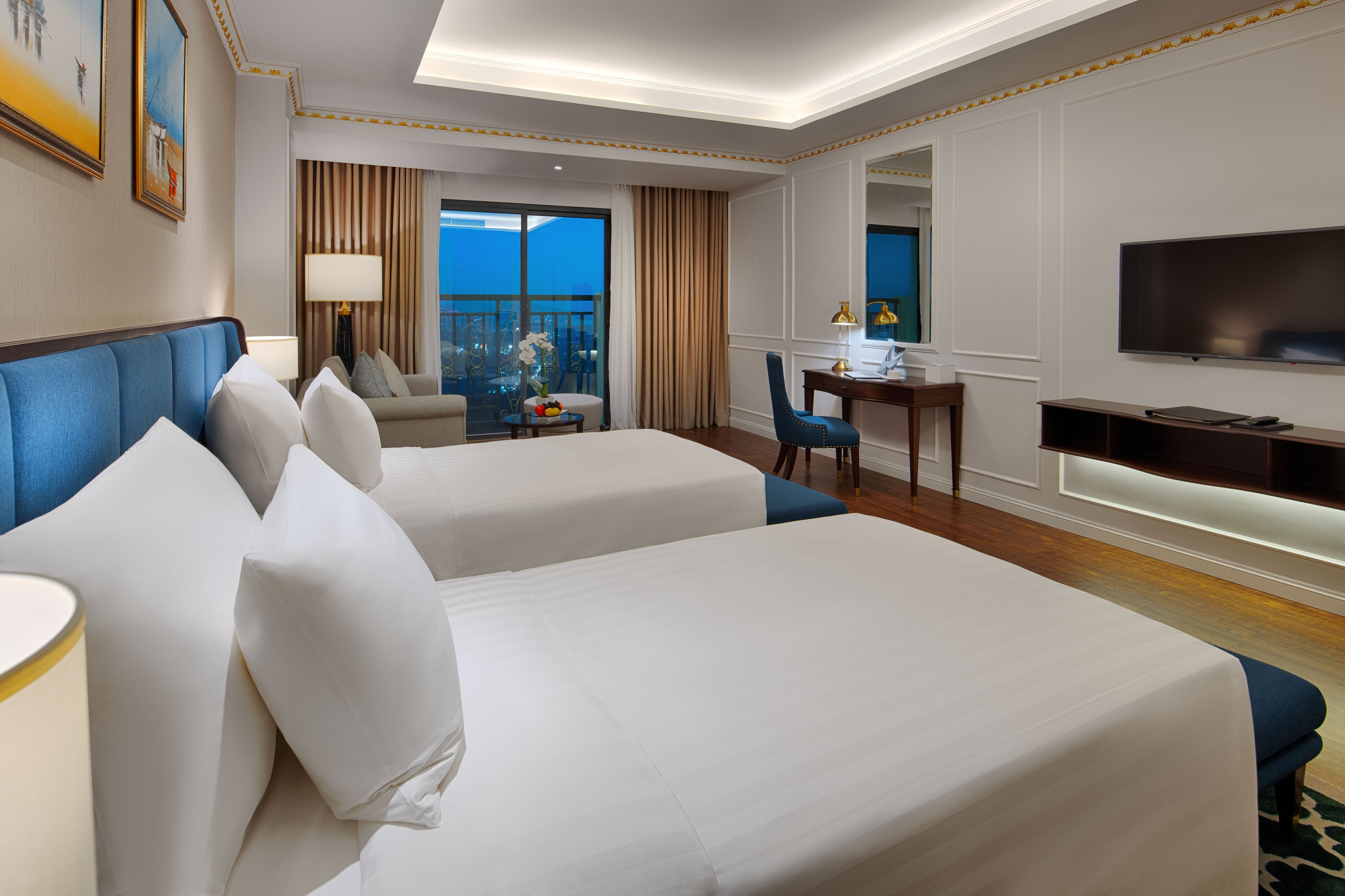 A hotel room with two beds, a desk, a chair, a TV, and a balcony with curtains.
