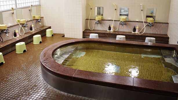 A public bath area with a central hot tub, multiple showerheads, and hand soap dispensers. - Business Ryokan Harada (Hiroshima)