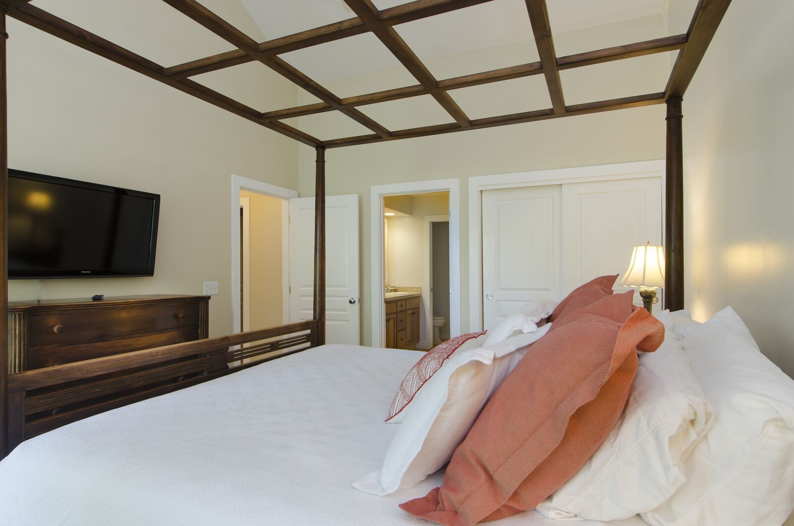 Un dormitorio con una cama grande, cabecera de madera, televisor montado en la pared, cómoda y una lámpara en la mesita de noche.