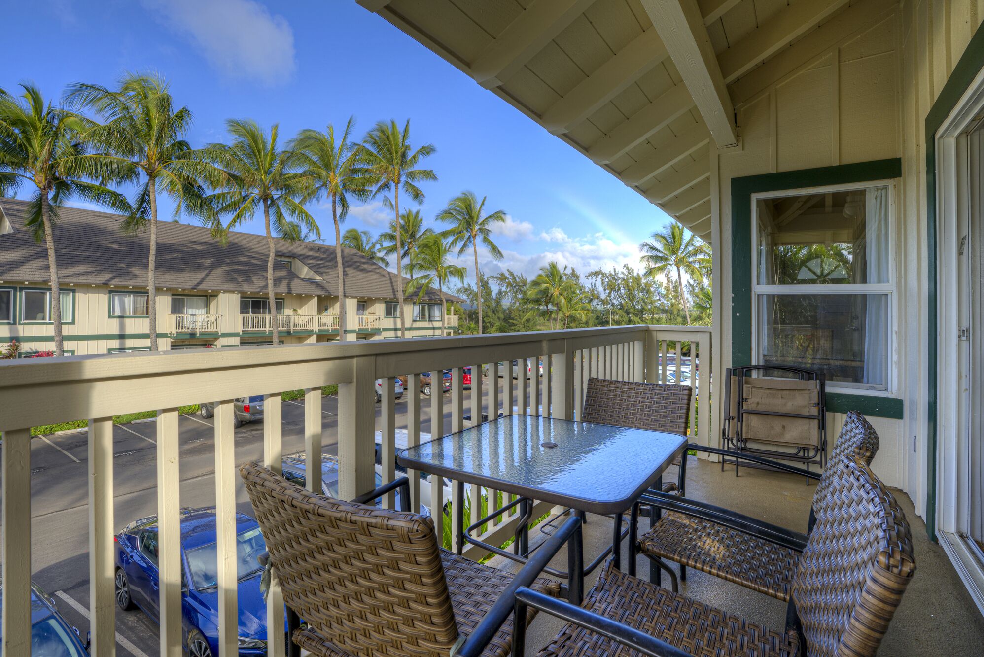 A balcony with wicker furniture, a glass table, and a view of palm trees and a building in the background.
