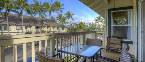A balcony with wicker furniture, a glass table, and a view of palm trees and a building in the background.