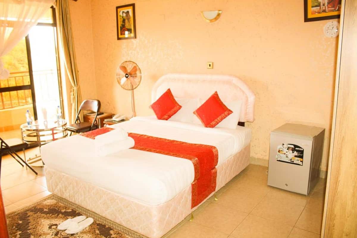 Habitación de hotel con cama, escritorio, silla, ventilador, mueble bajo y ventana con cortinas.