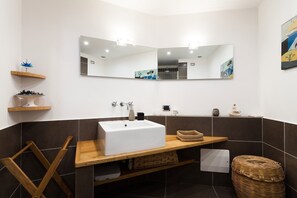 A modern bathroom with a white sink, wooden vanity, and a large mirror. There are shelves with decorative items and a wicker basket on the vanity. - Casa Rossella - My Rental Homes (Sorrento)
