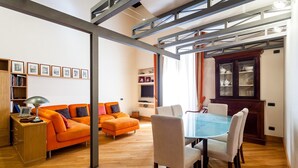 A modern living room with a wooden floor, orange sofa, white chairs, a glass dining table, and a bookshelf with framed pictures. - Casa Rossella - My Rental Homes (Sorrento)