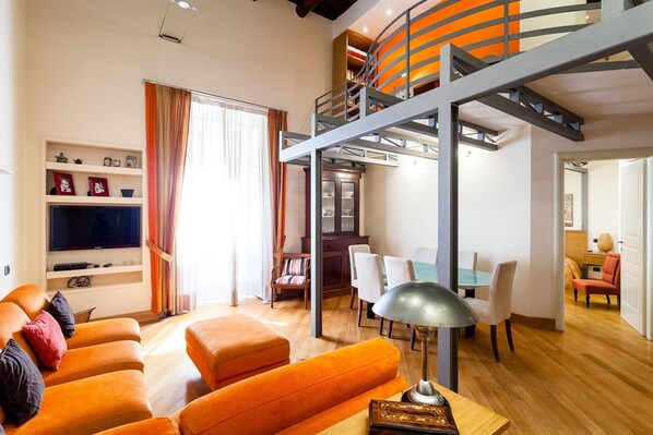 A modern living room with a large orange sofa, a flat-screen TV, and a dining area with a glass table. - Casa Rossella - My Rental Homes (Sorrento)