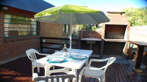 Un patio al aire libre con una mesa y sillas bajo un paraguas verde, una zona de barbacoa de ladrillo y un muro de ladrillo.