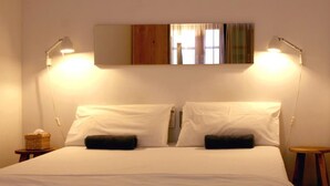 Una cama bien hecha con dos almohadas, una lámpara de noche y una mesita con una cesta.
