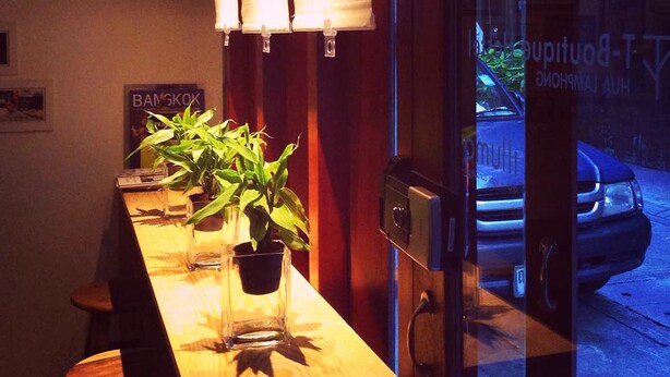 Una mesa de bar de madera con una planta en maceta, un letrero que dice "Bangkok" y una vista de la calle con un coche.