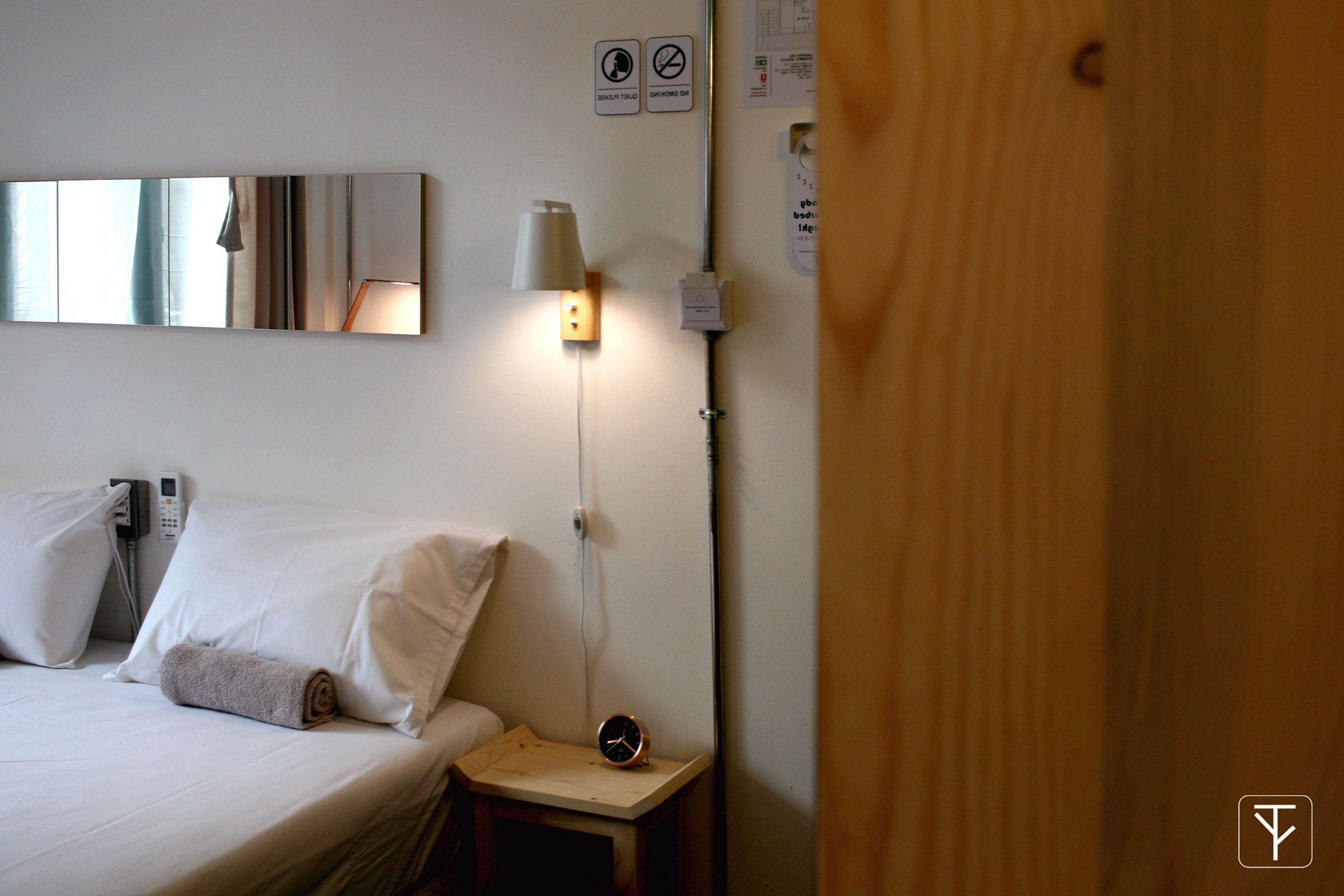 A hotel room with a bed, a bedside table with a lamp, a clock, and a mirror on the wall.