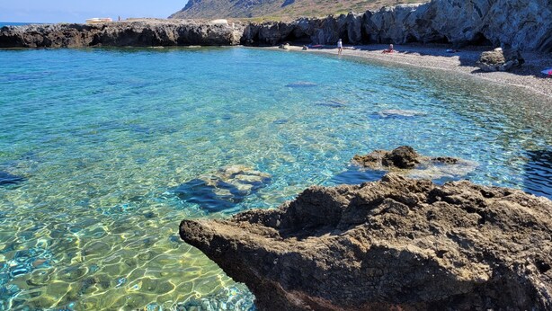 Una costa rocosa con aguas claras, una playa y una montaña al fondo.