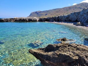 Eine felsige Küstenlinie mit klarem Wasser, ein Strand und ein Berg im Hintergrund.