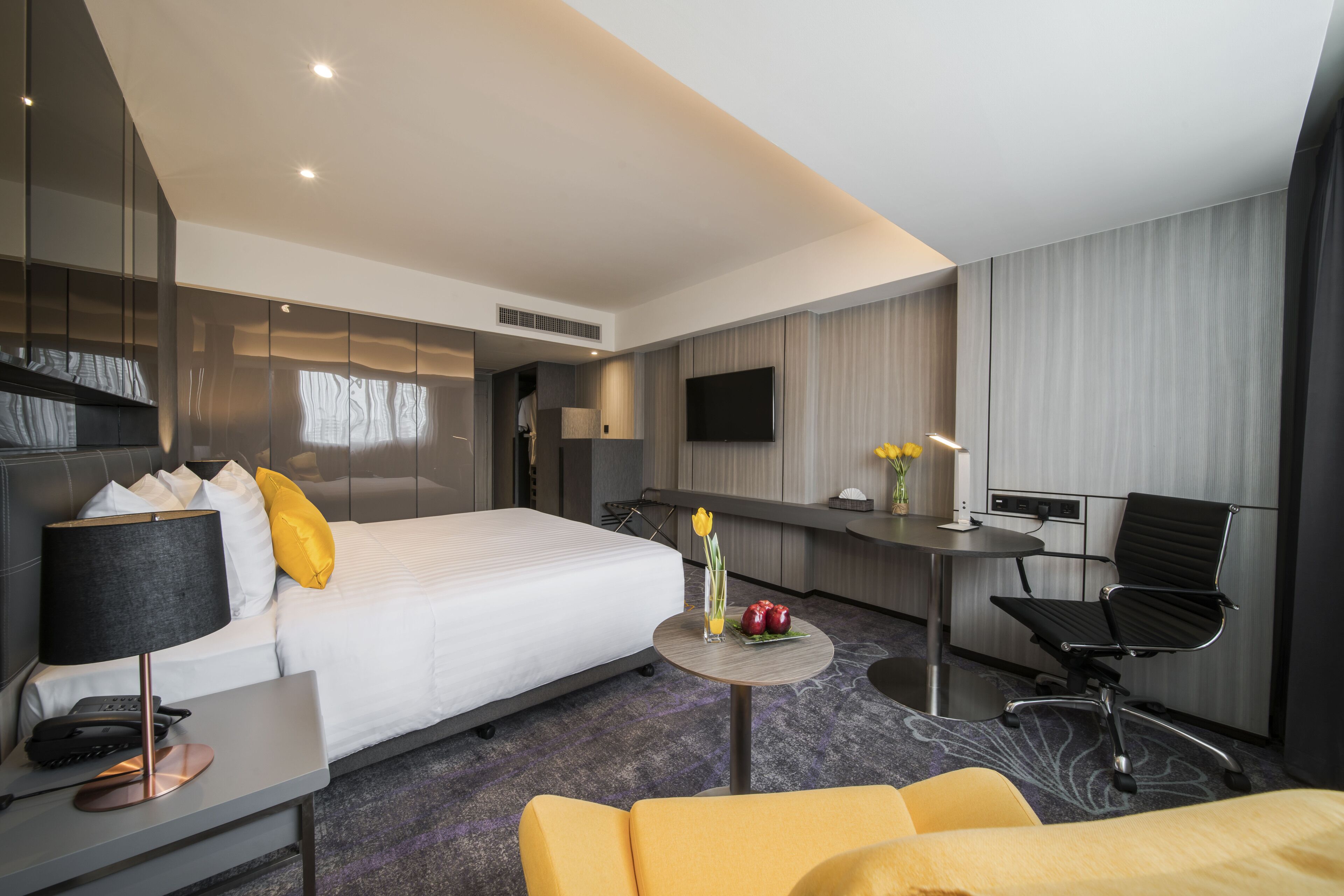 A modern hotel room with a large bed, a desk with a lamp, a chair, a small table with flowers, and a TV mounted on the wall.