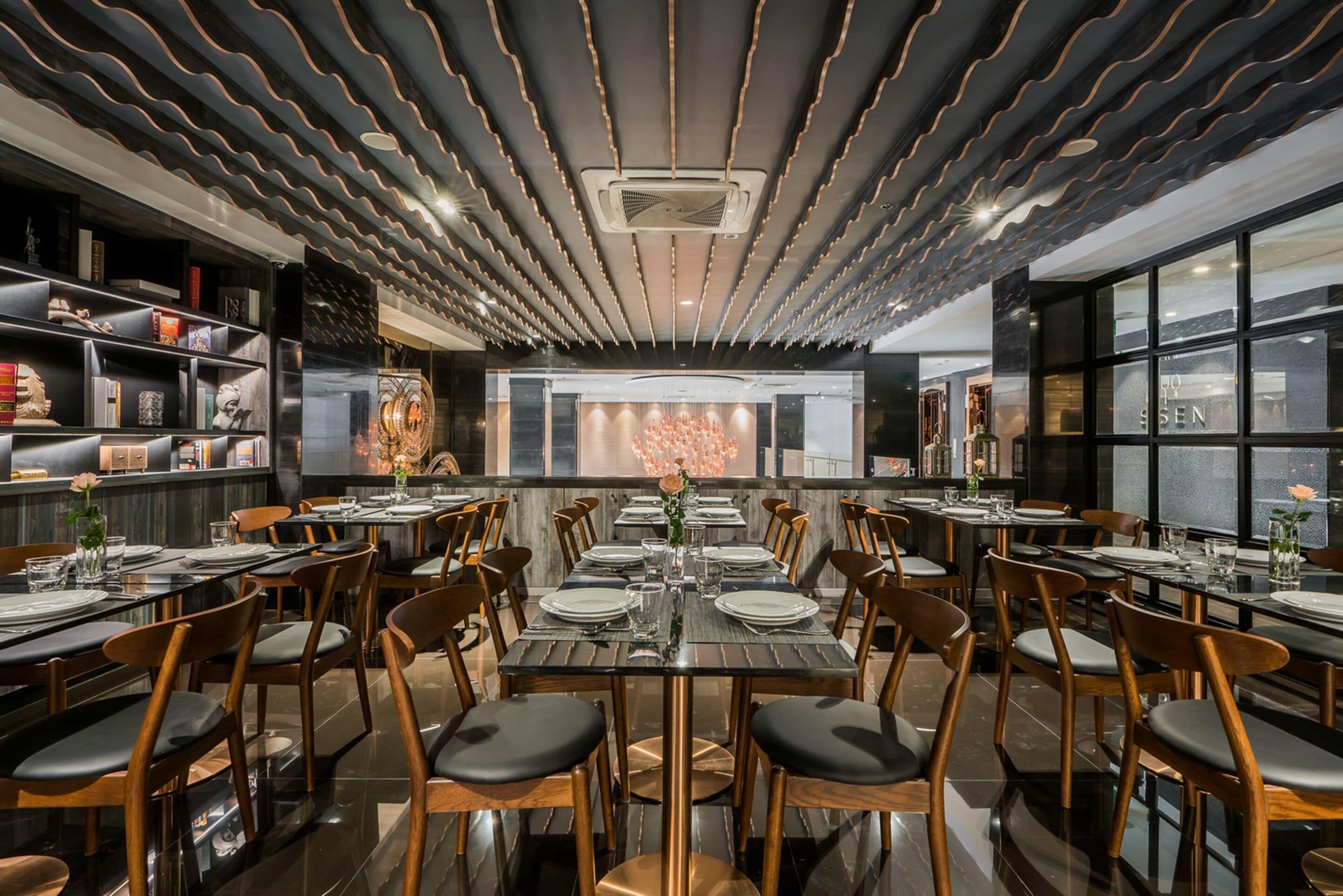 Un restaurante moderno con un área de comedor central que presenta una larga mesa rodeada de sillas, una pared decorativa con un arreglo floral y un techo con motivos.