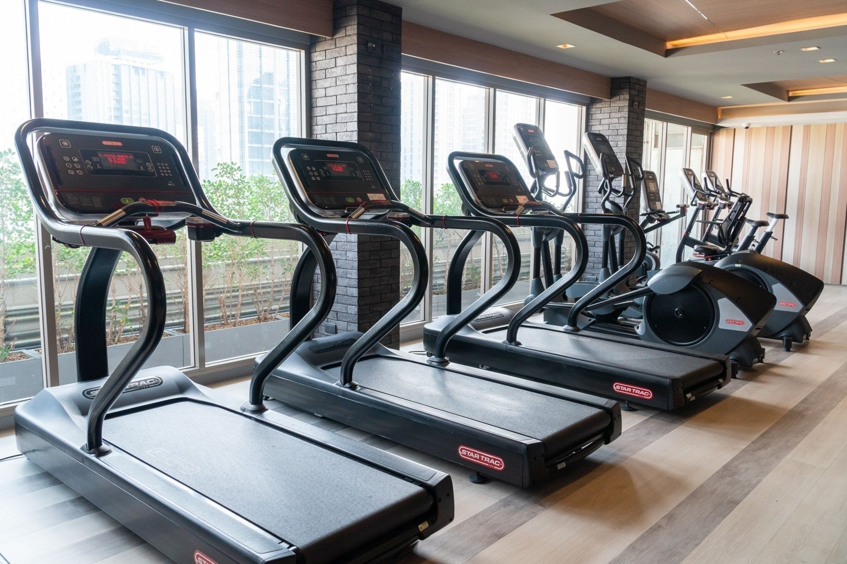 A row of treadmills and elliptical machines in a gym with large windows and a brick wall.