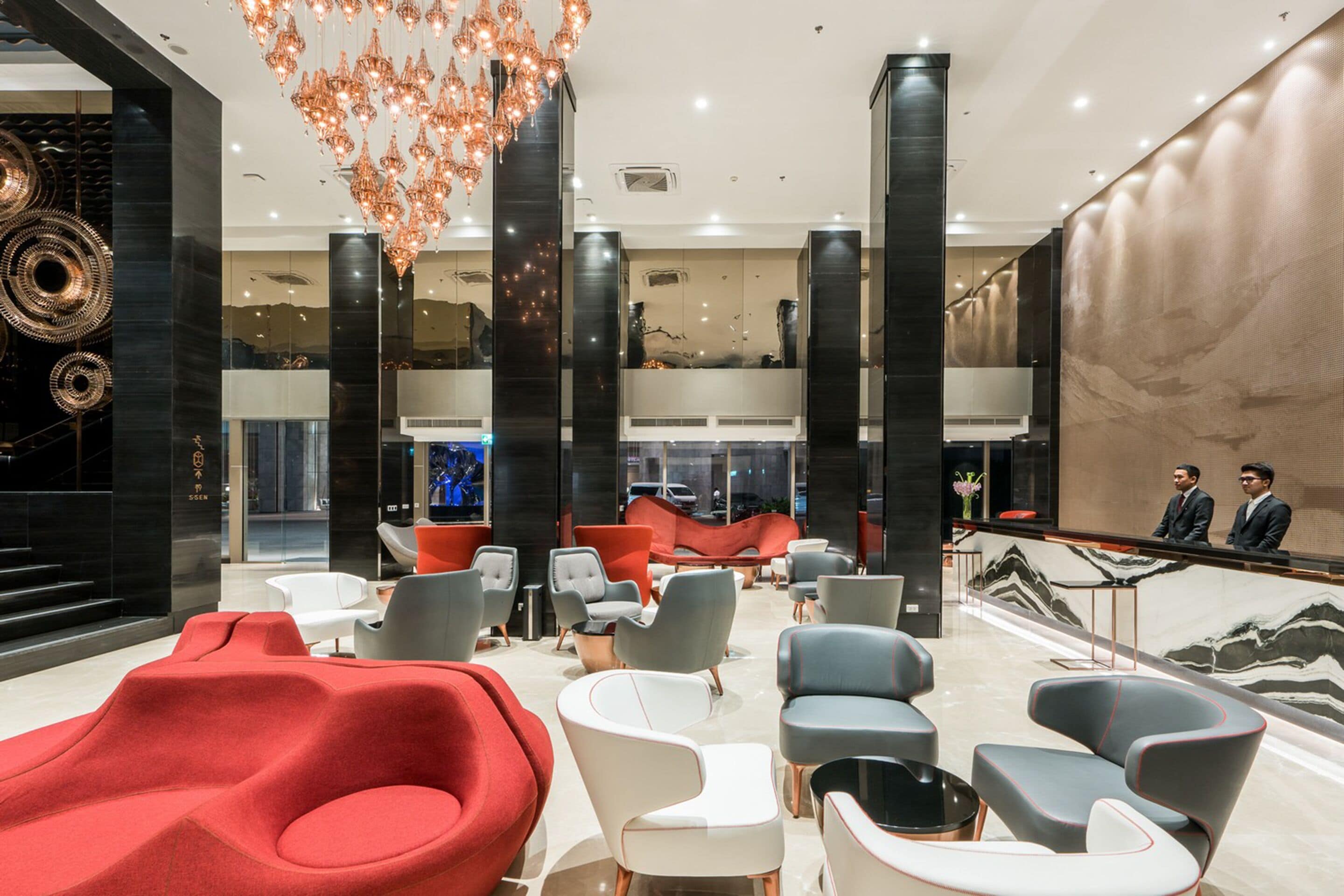 En modern entréhall med en blandning av röda och grå sittmöbler, en stor kristallkrona och en receptionsdisk.