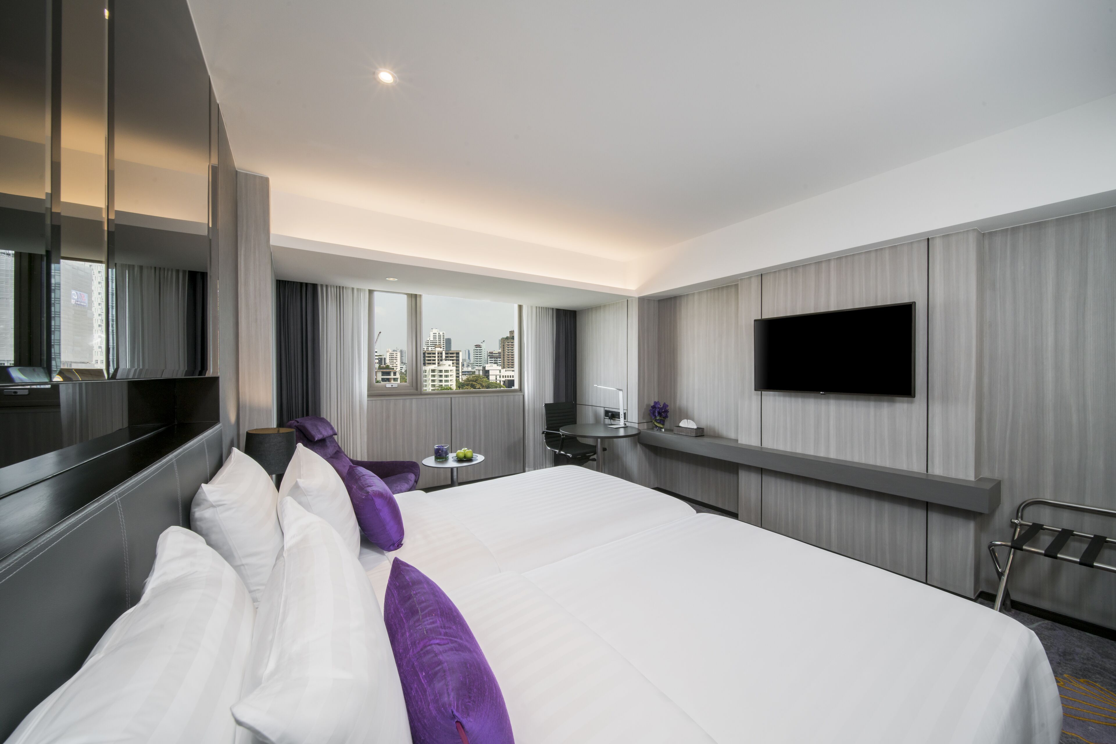 Una habitación de hotel moderna con una cama grande, un televisor de pantalla plana montado en la pared, una mesita con una lámpara y vistas panorámicas de la ciudad a través de amplios ventanales.