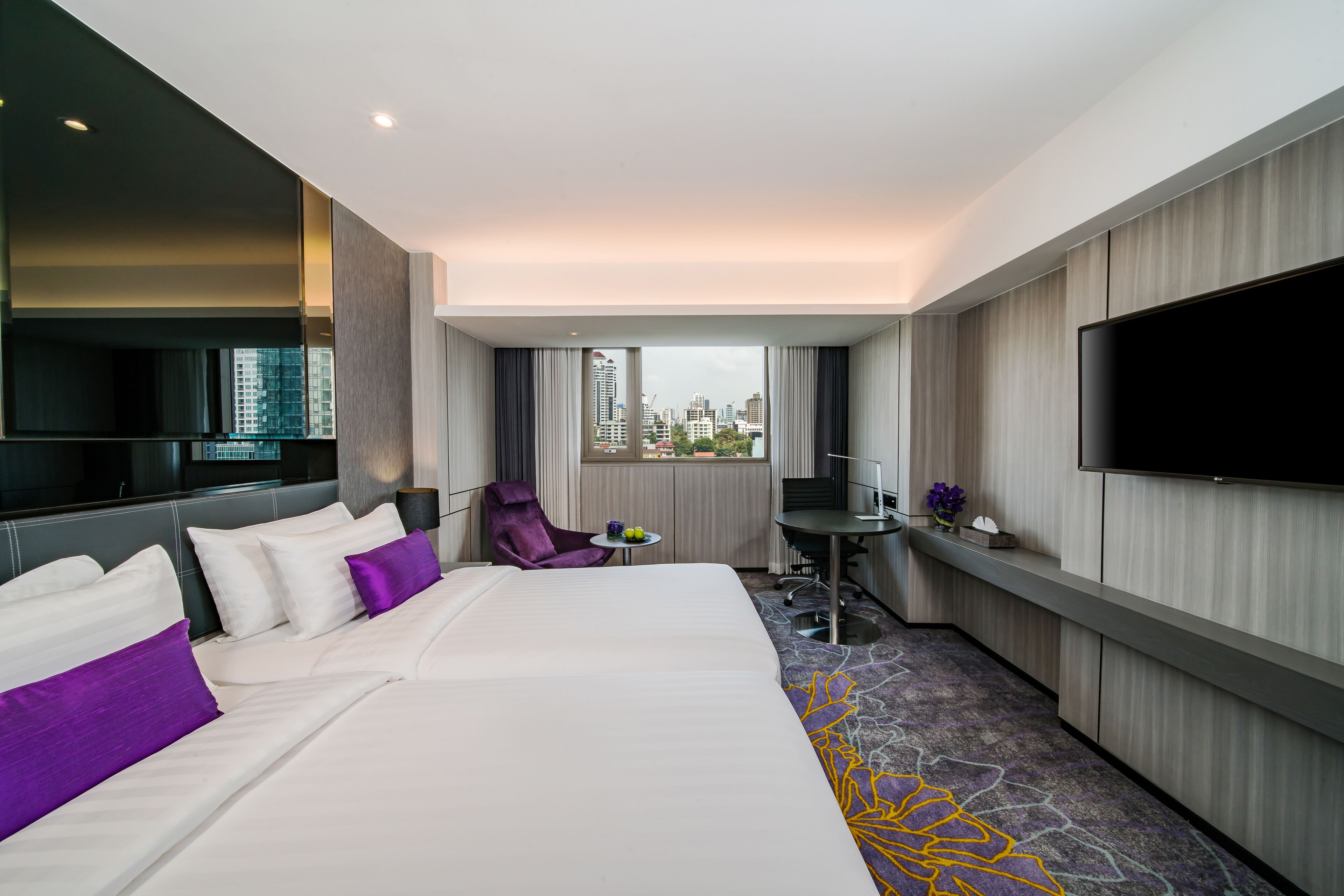 A hotel room with a large bed, a desk, a chair, a TV, and a view of the city.