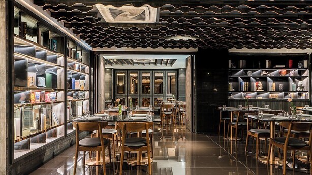 Een restaurant met een modern interieur, met een centrale eetruimte met tafels en stoelen, omringd door boekenkasten vol boeken en decoratieve objecten.