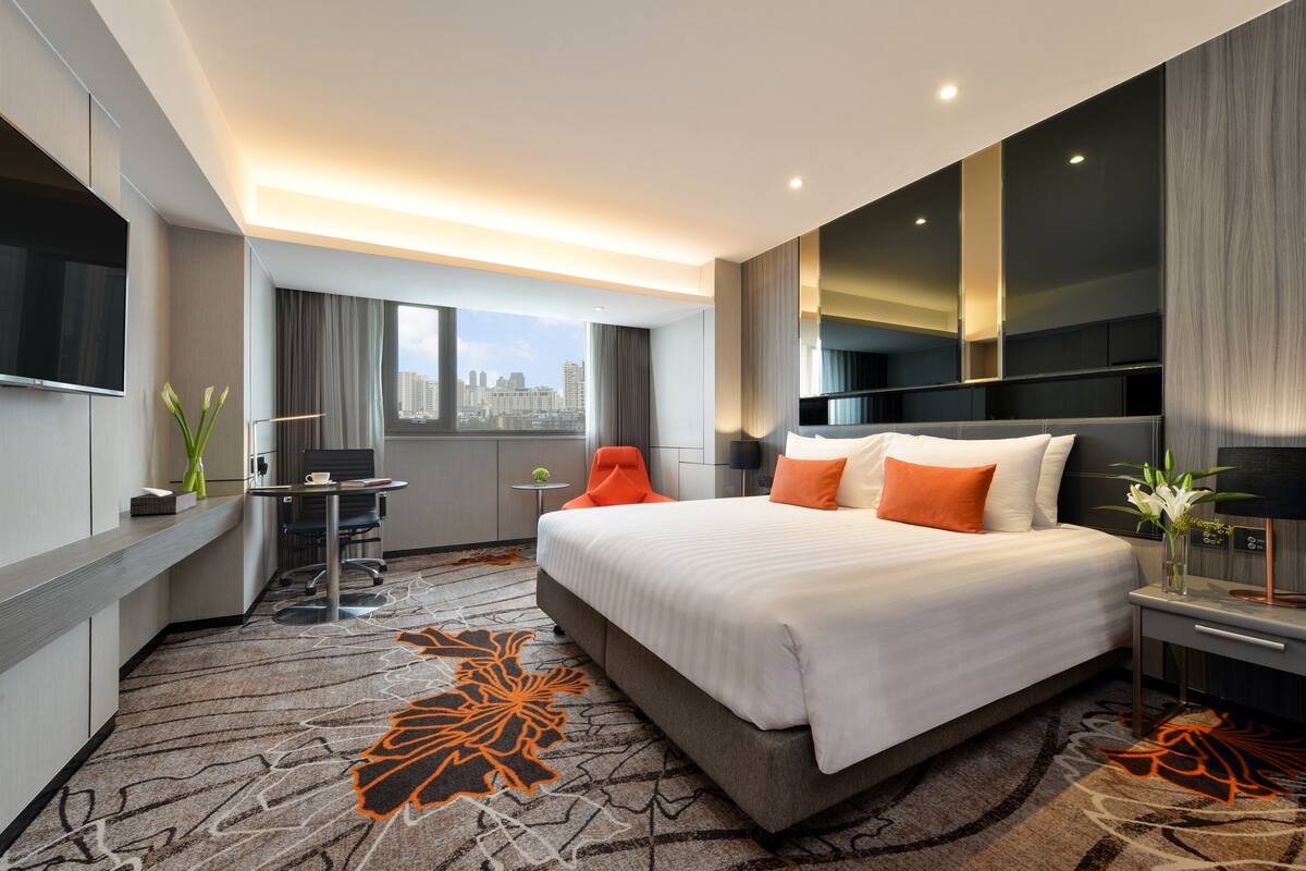 Ein modernes Hotelzimmer mit einem großen Bett, einem Schreibtisch, einem Sessel und Blick auf die Stadtlandschaft.