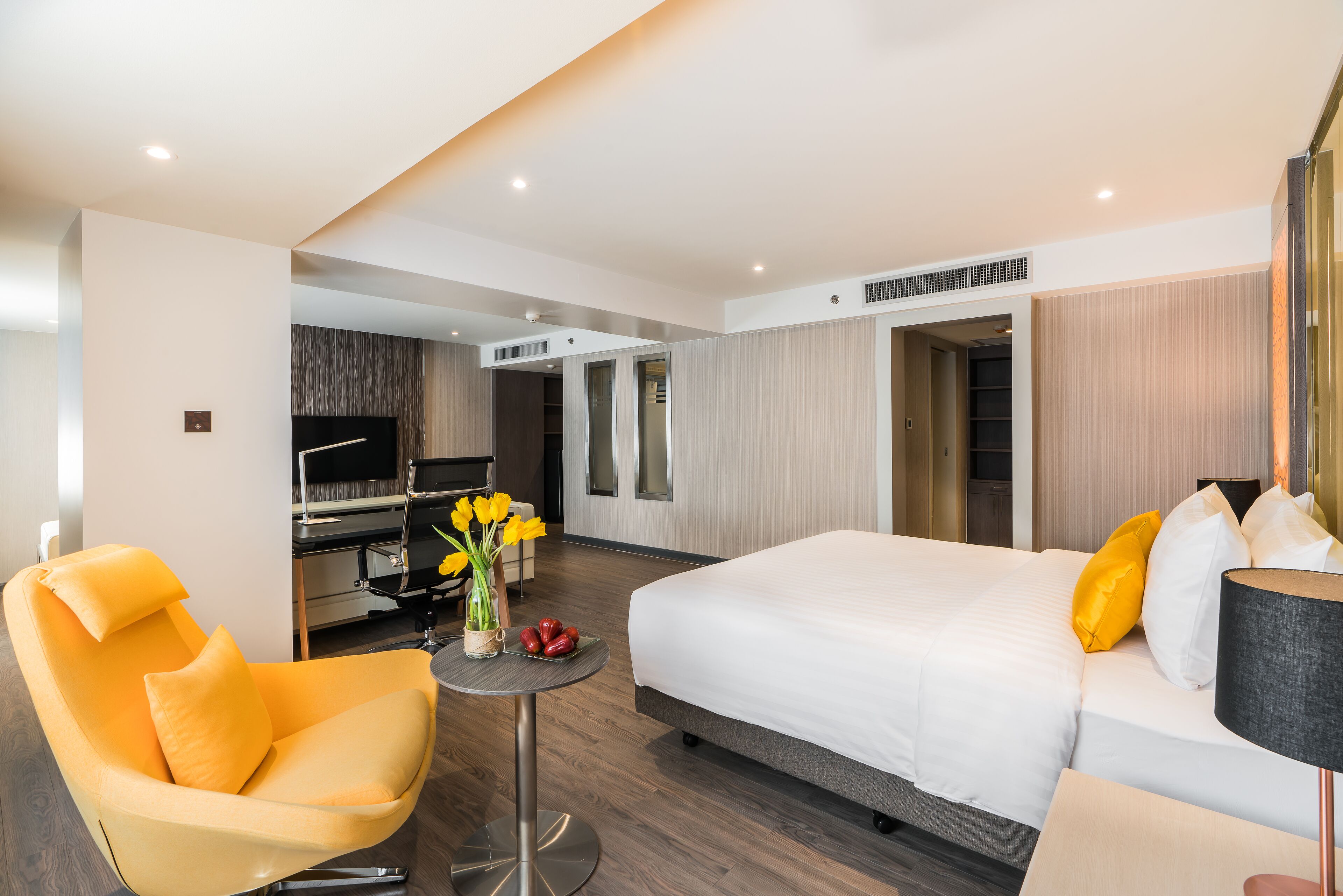 Een moderne hotelkamer met een groot bed, een geel fauteuil, een klein rond tafeltje met een bloemenvaas en een bureau met een computer en een televisie.