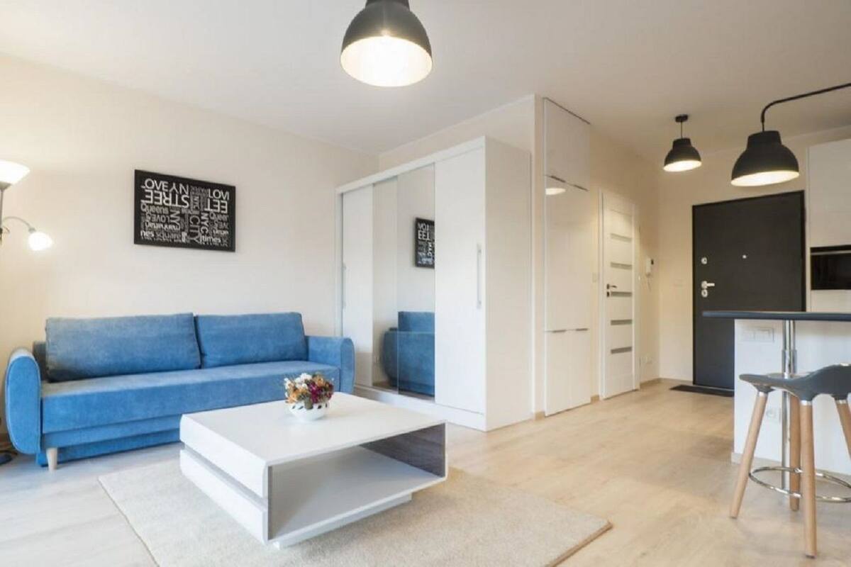 A modern living room with a blue sofa, a white coffee table, and black pendant lights. There is a kitchen area with a black countertop and white cabinets.