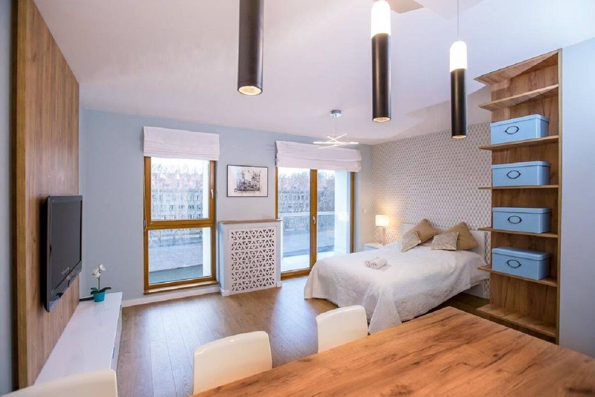A modern bedroom with a large bed, bedside tables, a desk, and a TV. There are windows with white curtains and a view of the outside.