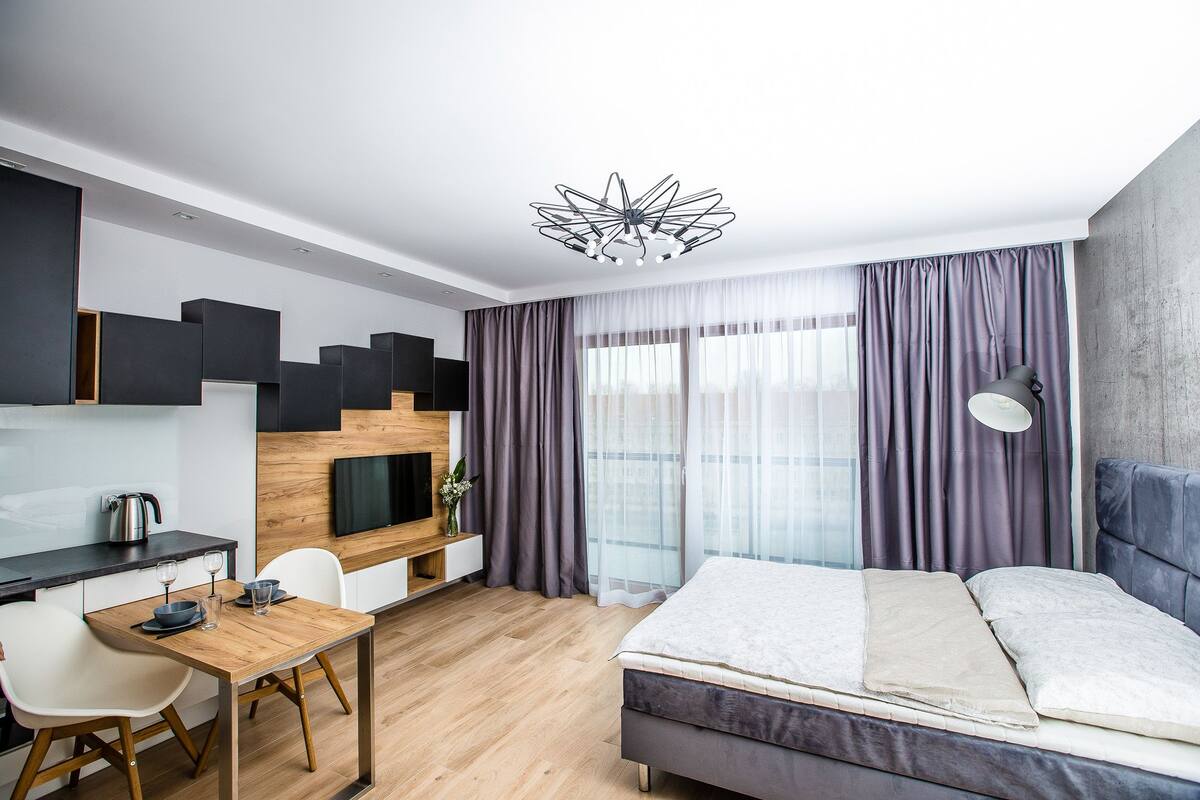 A modern hotel room with a bed, a desk with a TV, a dining area, and large windows with curtains.
