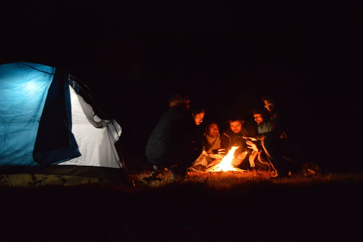 Um grupo de pessoas reunido em volta de uma fogueira em uma área coberta por tendas à noite.