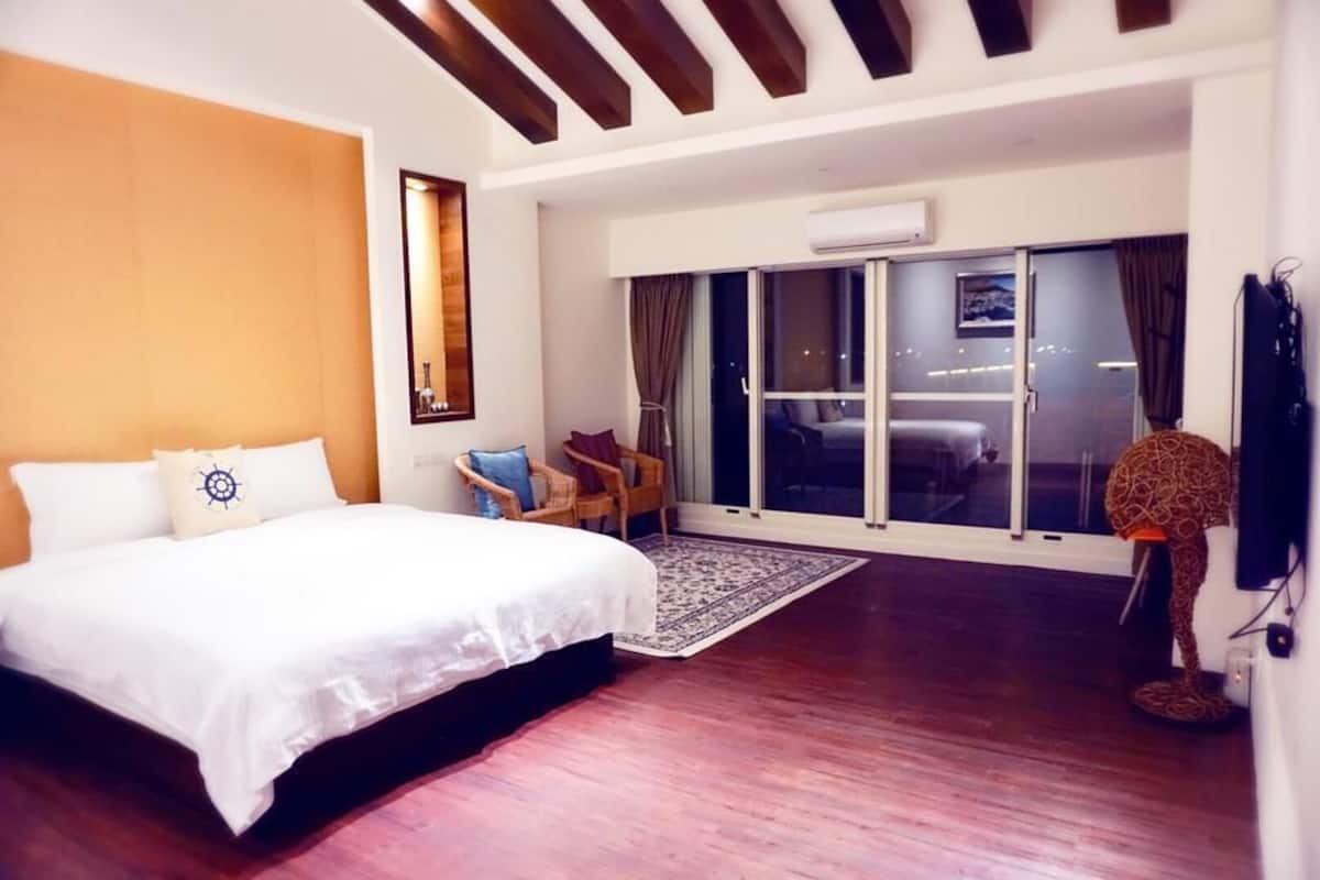 Een slaapkamer met een groot bed, een houten vloer, een raam met gordijnen, een stoel en een vloerlamp.