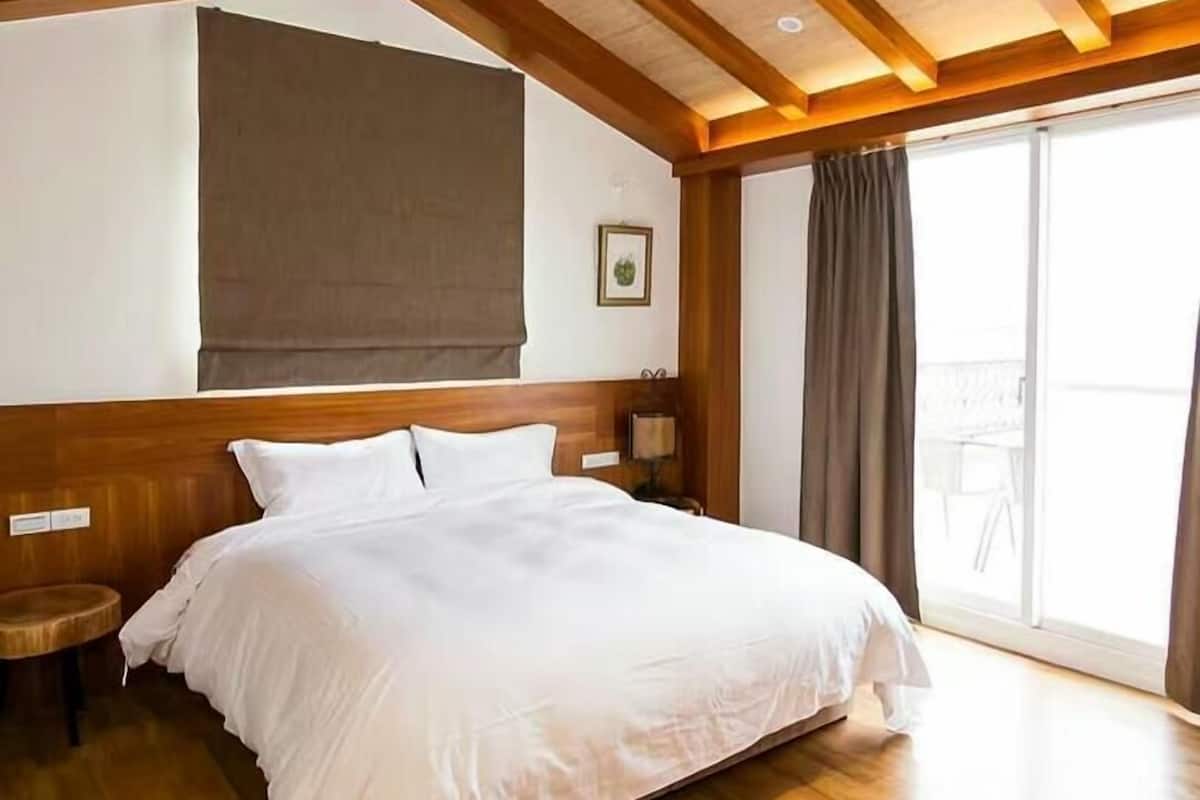 Een hotelkamer met een groot bed, houten balken in het plafond, een raam met gordijnen en een klein nachtkastje.