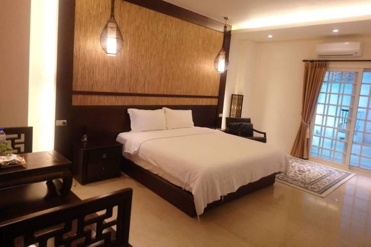 Een slaapkamer met een groot bed, nachtkastjes, een bureau en een stoel. Er is een raam met gordijnen en een airconditioning aan de muur.