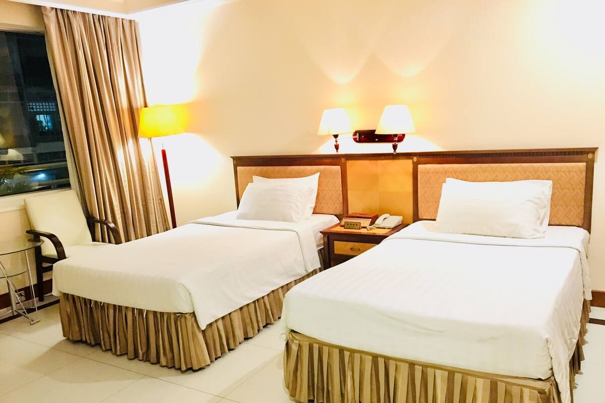 A hotel room with two beds, a nightstand with a phone, a lamp, and a view of the outside through a window with curtains.