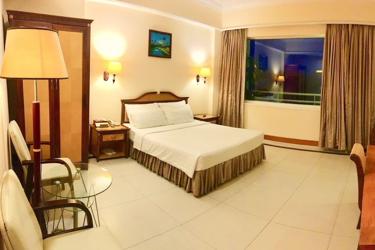 A hotel room with a bed, bedside tables, a desk with a tray of breakfast items, a chair, a lamp, and a painting on the wall.