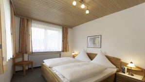 A double bed with white linens, a wooden nightstand with a lamp, a chair, a window with curtains, and a framed picture on the wall. - Ferienwohnungen Wölfl (Bodenmais)