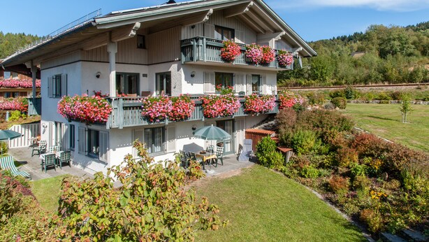 A two-story building with balconies adorned with flowers, a patio area with outdoor seating, and a well-maintained garden. - Ferienwohnungen Wölfl (Bodenmais)