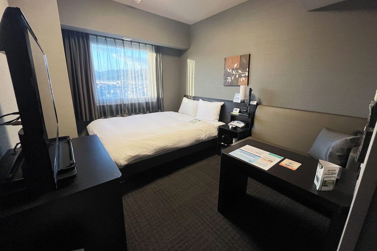 Een hotelkamer met een bed, nachtkastje, lamp en uitzicht op de stad door het raam.