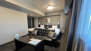 A hotel room with a bed, a black leather sofa, a small table with a remote control, and a window with curtains.