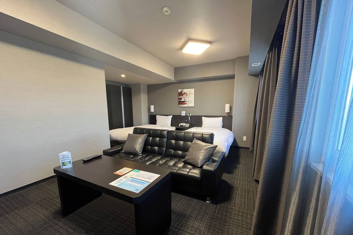 Een hotelkamer met een bed, een zwarte leren bank, een klein tafeltje met een afstandsbediening en een raam met gordijnen.