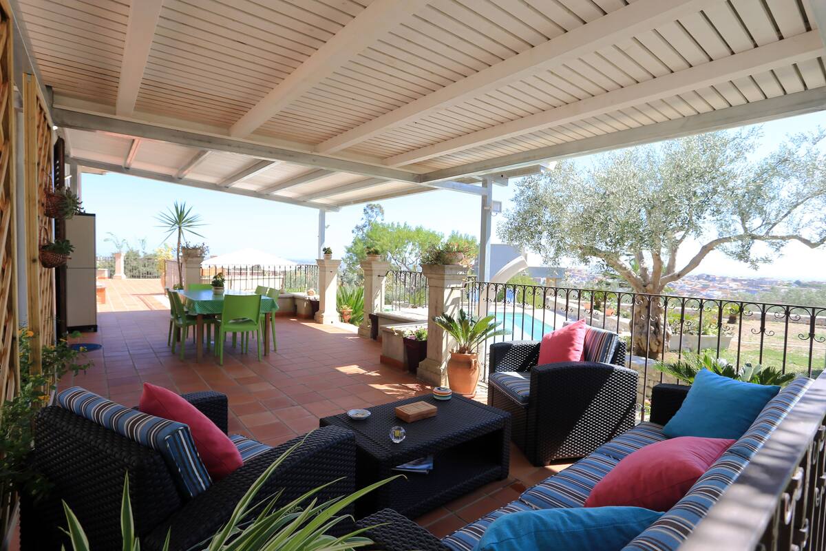 Un patio exterior con mobiliario de mimbre, una mesa de comedor y sillas. Se observa una piscina al fondo.