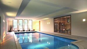 A large indoor swimming pool with clear blue water, surrounded by a tiled floor and a wooden deck. There are lounge chairs and a table near the pool, and a large window on one side. - Seehotel am Neuklostersee (Zurow)