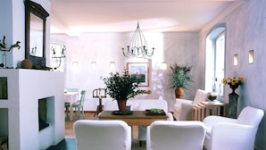 A spacious living room with a wooden dining table, white upholstered chairs, a chandelier, and a large potted plant. - Seehotel am Neuklostersee (Zurow)