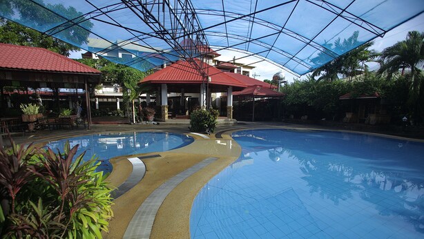 An indoor swimming pool with a curved design, surrounded by potted plants and a covered area with a red roof. - Villa Elita (Sudipen)