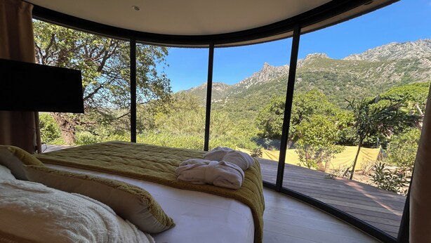 Quarto com uma janela grande, uma cama com almofadas e vista para montanhas.
