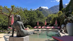 Estátua de uma figura sentada perto de uma piscina com espreguiçadeiras e guarda-sóis, rodeada por vegetação e montanhas.