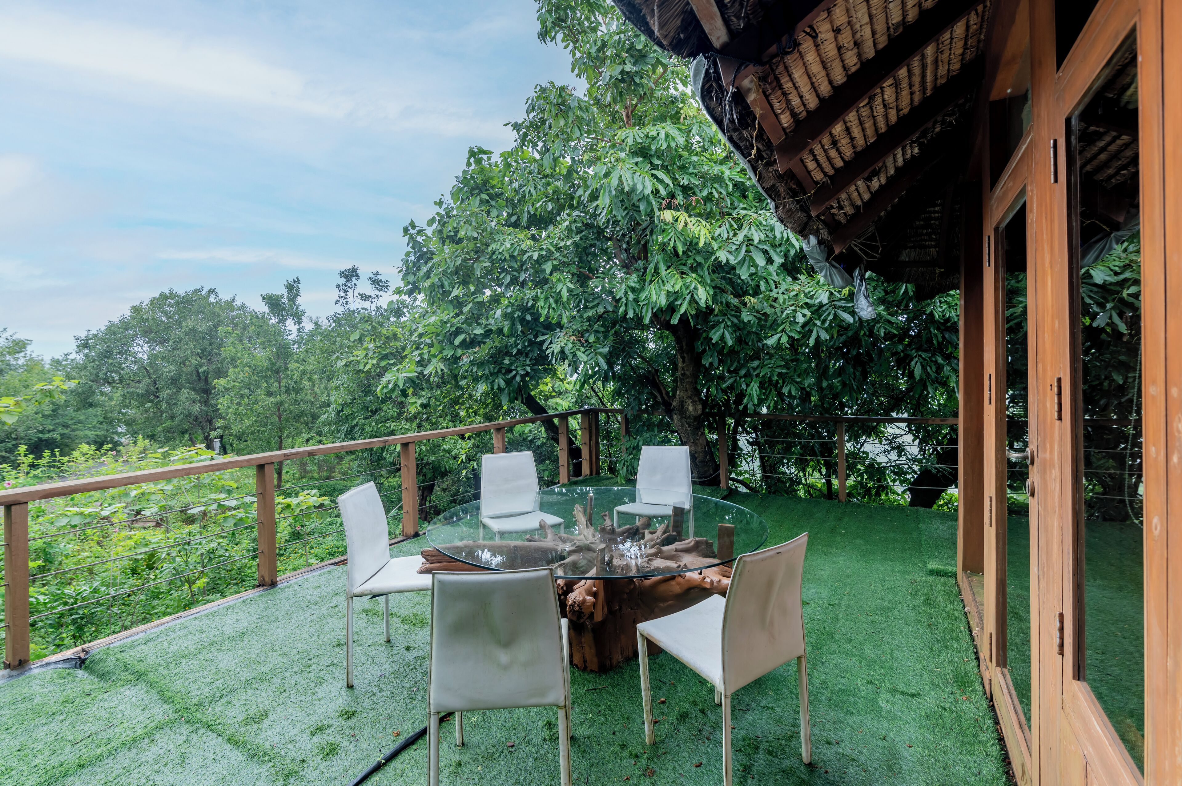 A glass-top table with four chairs on a green artificial lawn, surrounded by wooden railings and trees.