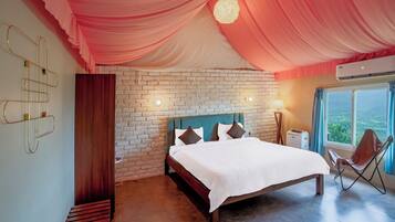 A bedroom with a brick wall, a bed with white bedding, a blue headboard, and a view of greenery through a window.